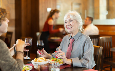 Senior woman in cafe sits opposite male colleague in cafe, chat and joke, drinking wine, spend time. Date, romantic meeting, nice time together. Atmosphere of cafe, restaurant conducive communication