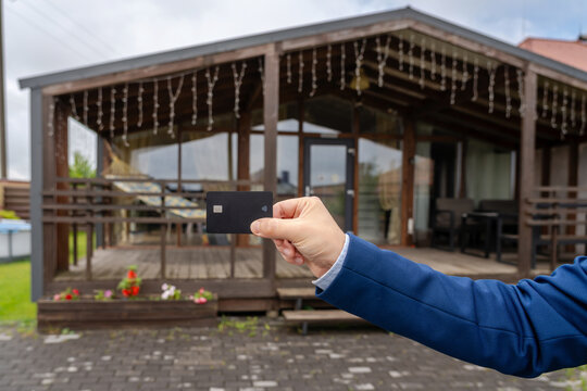 Plastic credit card in hand with new house at background. Broker, mortgage, property, real estate concept. Buying or renting