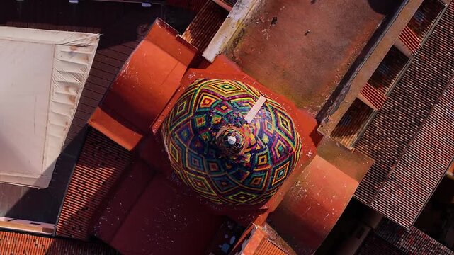 Aerial view of a vibrant, geometrically patterned dome contrasts sharply with the surrounding red-tiled rooftops, Sardinia, italy.