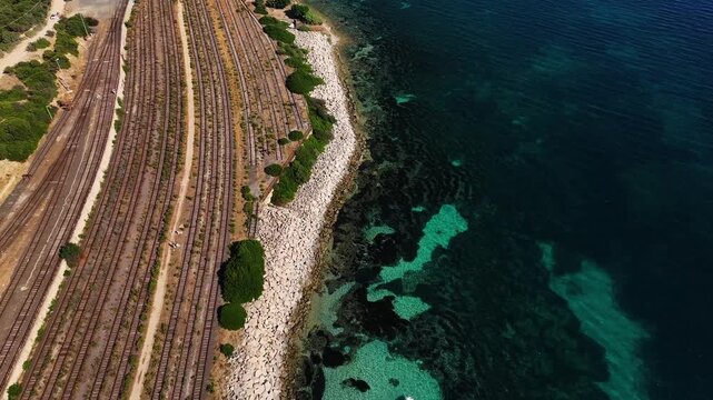 Aerial view of railway tracks running parallel to the rocky coastline, contrasting with the clear turquoise waters of the sea, Sardinia, italy.