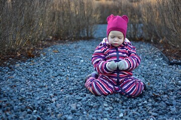 Little girl in a striped winter suit sitting on gravel and holding a small stick while exploring outdoors. Candid moment of childhood discovery in nature.