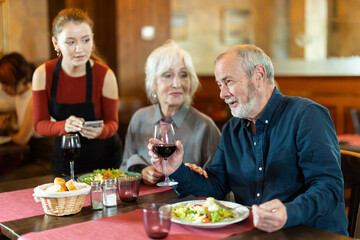 Positive female server listening carefully while standing beside old couple enjoying evening meal in restaurant