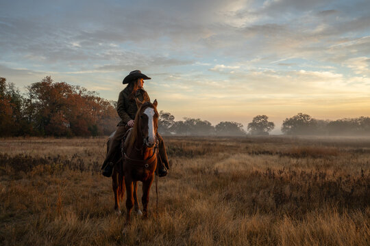 Woman Wearing Cowboy Hat Riding Horse and Looking Off At Golden Hour of Sunset With Cloudy Sky
