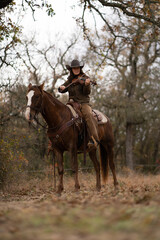 Cowgirl Woman Wearing Cowboy Hat Looking Down and Playing Fiddle Violin While Sitting On Brown and White Horse In Front of Trees and Cloudy Sky