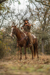 Cowgirl Woman Wearing Cowboy Hat Looking Down and Playing Fiddle Violin While Sitting On Brown and White Horse In Front of Trees and Cloudy Sky