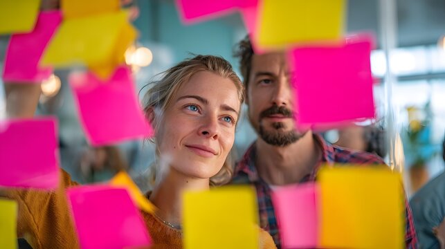 Man and a woman are looking at a wall covered in yellow and pink sticky notes. The woman is smiling and the man is looking at her
