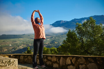 Naklejka premium Woman In Hijab Stretches Arms Amid Mountain Landscape, Outdoor Fitness Moment