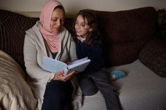 Mother in Hijab Reading a Book to Daughter on Sofa at Home