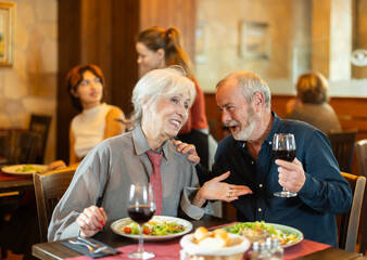 Happy senior couple enjoying relaxed dinner together in cozy restaurant while talking and sharing wine