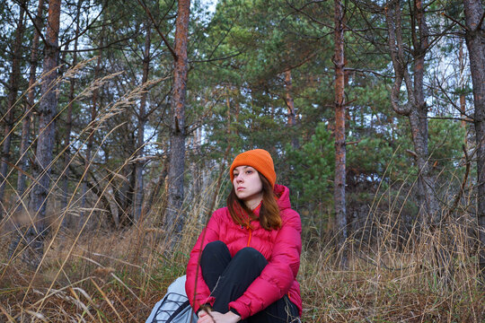 A young woman is sitting on the grass in the autumn forest and looking into the distance. Resting, hiking in the forest, hiker at rest