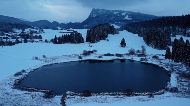 Aerial view of the dark, still waters of Lac Ter surrounded by a snow-covered landscape and forests under a cloudy sky, Lake Ter, Vaud, Switzerland.
