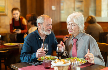 Senior spouses in cafe, chat and joke, flirting, eat and drink wine, spend time. Date, romantic meeting, nice time together. Atmosphere of cafe, restaurant conducive communication