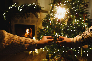 Hands holding fireworks against christmas tree lights in evening cozy living room. Happy New Year eve! Atmospheric family celebration with burning sparklers in hands close up