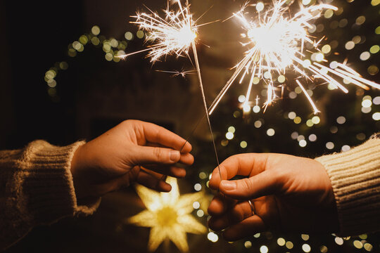 Happy New Year! Burning sparklers in hands on background of christmas tree golden lights bokeh at night. Glowing fireworks in couple hands, new year eve holiday celebration - Powered by Adobe