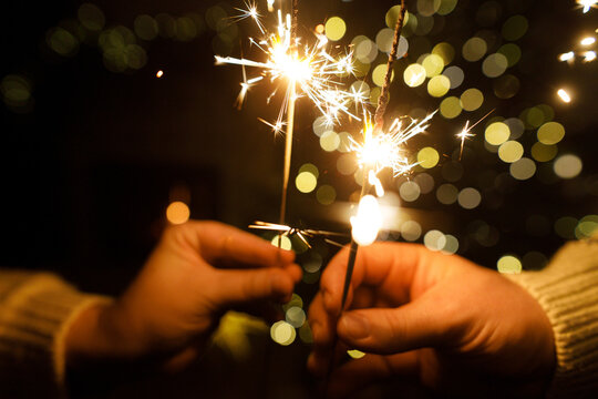 Hands holding fireworks against christmas tree lights bokeh in evening cozy living room. Happy New Year eve! Atmospheric family celebration with burning sparklers in hands close up - Powered by Adobe