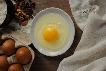 A wooden table with baking ingredients, including a cracked egg in a white bowl, flour, chocolate chips, walnuts and almonds, as well as brown eggs in a carton, ready for homemade desserts.