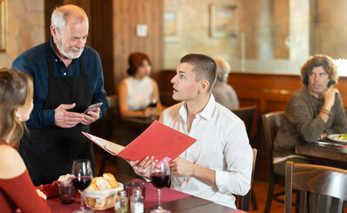 Fototapeta premium At cafe hall, senior man waiter communicates with girl and boyfriend customers, take order, answer questions about composition of dish of day. Client consult with waiter about vegan health menu .