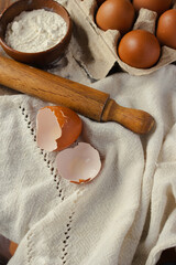 Vertical top view of freshly baked ingredients, a brown eggshell next to other whole eggs in a carton, bowls of flour and a rolling pin, ready to make homemade cakes and healthy recipes