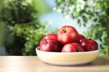 Ripe red apples in bowl on wooden table against blurred green background, closeup. Space for text
