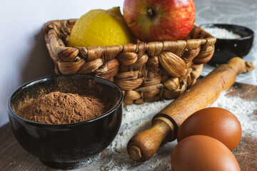 Baking ingredients on a rustic wooden surface with flour dusting, bowl of cocoa, eggs, rolling pin and a woven basket holding an apple and a lemon for homemade recipes