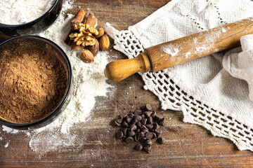 A selection of delicious ingredients for gourmet chocolate cakes and desserts, including flour, cocoa powder, nuts, chocolate pieces and a rolling pin, all presented on a rustic wooden board.