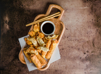 Asian appetizers including a crispy egg roll, fried cream cheese wontons, and a pork dumpling (potsticker) served with soy sauce on a dark textured background.