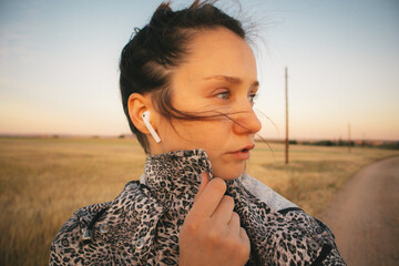 A young woman looking in a distance walking in a countryside path. Brunette girl is listening to music with headphones in windy day. Rural lifestyle.