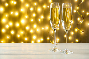 Merry Christmas and Happy New Year. Flute glasses with sparkling wine on white wooden table against golden background with serpentine streamers and blurred lights, closeup. Space for text