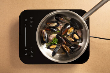 Fresh mussels are simmering in a stainless steel pot with herbs on an induction cooktop. The kitchen features a minimalist design, creating an efficient cooking space.