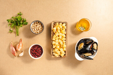 Fresh ingredients are organized on a light wooden table, including pasta, beans, mussels, and...