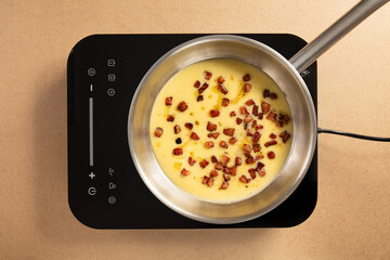 Savory dish being prepared with whisked eggs and crispy bacon bits on a sleek induction stove. A warm, inviting kitchen atmosphere enhances the cooking experience.