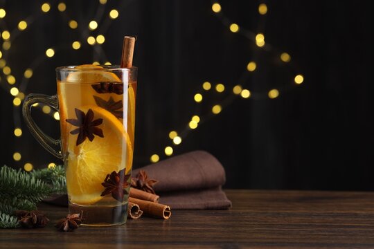 Delicious mulled wine with slices of orange, spices and fir branches on wooden table against black background with blurred Christmas lights, closeup with space for text. Bokeh effect
