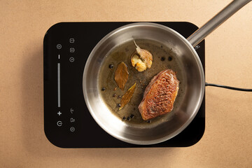 Seared steak cooking in a stainless-steel pan with garlic, bay leaves and peppercorns on a modern induction cooktop. Top-view shot of gourmet meat preparation.