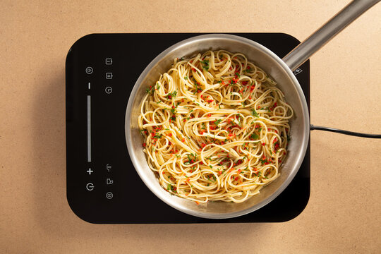 A pot of spaghetti noodles is tossed with garlic and herbs, placed on a sleek induction cooktop. The kitchen appears organized and well-designed, reflecting a modern aesthetic.
