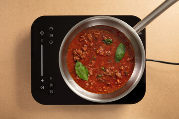 A rich meat sauce bubbles gently in a stainless steel pan with fresh basil leaves on top. This scene depicts a modern kitchen with a sleek induction cooktop ready for cooking.