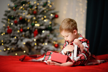 child in festive plaid pajamas opening a present while sitting near a beautifully lit Christmas tree. Warm holiday atmosphere with soft bokeh lights and cozy seasonal colors.