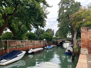 Fototapeta premium A narrow Venetian canal framed by lush trees and red brick walls. Several small boats are moored along the banks. In the distance, a small, traditional stone bridge.