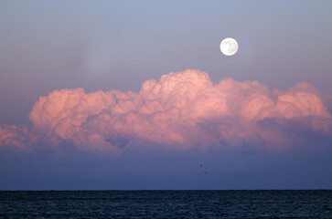 Full moon shining brightly over a calm ocean with pink and purple clouds at dusk