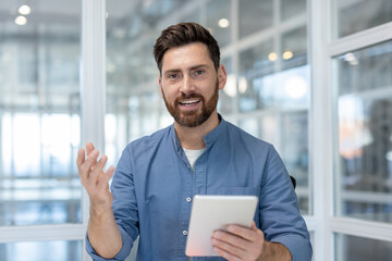 Smiling businessman in smart-casual attire holding a tablet and gesturing while presenting ideas during a virtual meeting or webinar in a modern office setting