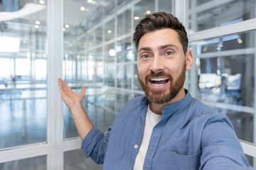 Happy entrepreneur vlogging from a bright modern glass office, smiling and pointing as he...