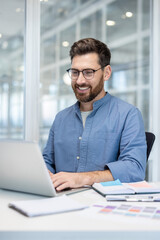 Bearded businessman in glasses smiling while typing at laptop in a modern office, confident and...