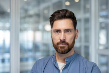 Obraz premium Confident man with a beard and serious expression looking at the camera, standing in a blurred modern office building, representing professionalism and business success