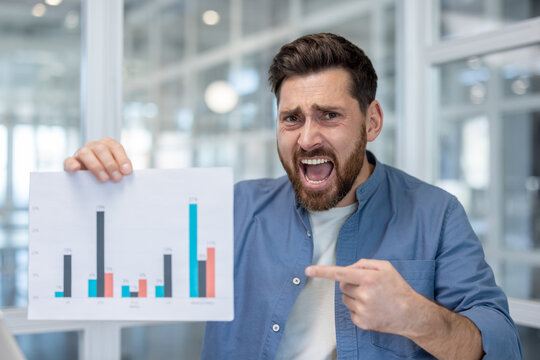 Stressed businessman in modern office angrily pointing at a bar chart with declining results, shouting in frustration over negative financial data and report failure