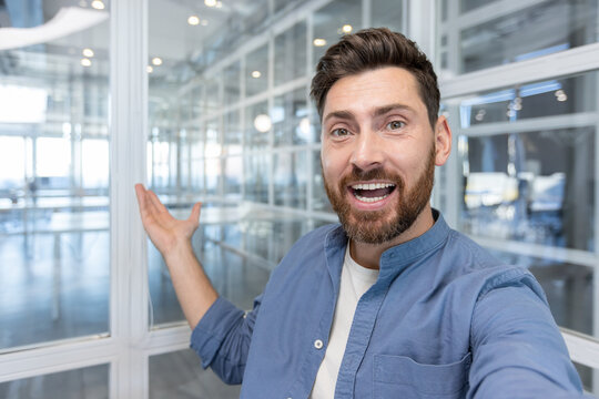 Happy entrepreneur vlogging from a bright modern glass office, smiling and pointing as he introduces the open workspace to his online audience with enthusiasm and confidence