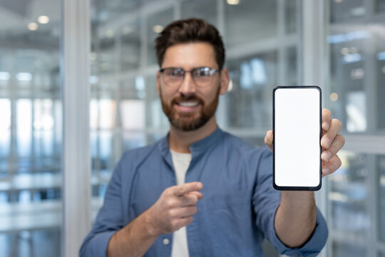Smiling man presenting and recommending mobile phone with empty white display template, offering space for advertising or app interface design in modern office