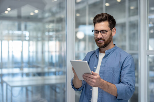 Smiling man in casual business attire stands in a modern office, using a tablet to manage work and communicate-confident, relaxed professional embracing mobile technology and productivity