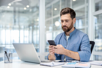 Businessman using smartphone for communication and productivity while working at a desk with a laptop and documents in a contemporary bright office environment