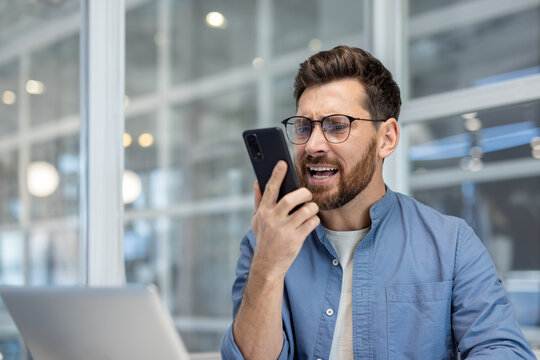 Bearded man in modern office yelling into his smartphone during a stressful work call, showing frustration, anger and conflict while handling remote work or business problems