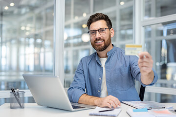 Fototapeta premium Smiling man at desk holding a credit card while using a laptop for an online payment, representing secure e-commerce, digital banking and convenient remote transactions in a modern office