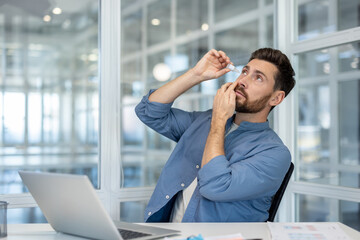 Man at a bright modern office desk applying eye drops to relieve dry eyes, emphasizing eye care, health, and discomfort experienced while working on a computer